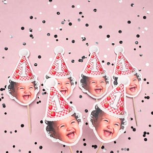 May include: Six cupcake toppers featuring a smiling child's face wearing a red and white gingham party hat with a white pom-pom on top. Each hat displays a strawberry pattern and the number one in white. Each topper is attached to a wooden stick.