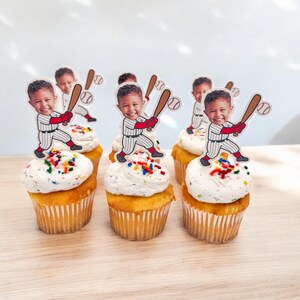 May include: Cupcakes topped with white frosting and colorful sprinkles. Each cupcake has a baseball-themed topper featuring a smiling child in a baseball uniform holding a bat. The cupcakes are in paper liners.