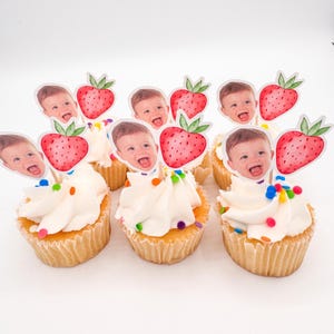 May include: Cupcakes with white frosting and colorful sprinkles. Each cupcake is topped with a paper cutout of a baby's face and a red strawberry. The cupcakes are arranged on a white surface.
