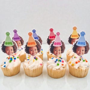 Puede incluir: Cupcakes con glaseado blanco y chispas de colores. Cada cupcake tiene una foto de un ni&ntilde;o con un gorro de fiesta que dice "ONE". Los gorros son de varios colores, incluyendo verde, morado, naranja, azul, rosa y amarillo. Los cupcakes est&aacute;n en moldes de papel.