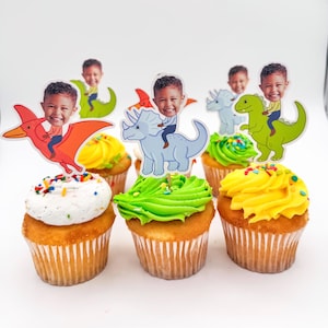 May include: Six cupcakes with colorful frosting and dinosaur-themed toppers. Each cupcake has a different color frosting, including white, yellow, and green. The toppers feature a child's face riding various dinosaurs.