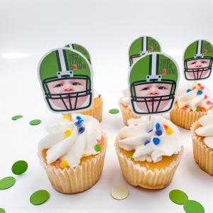 May include: Cupcakes with white frosting and colorful sprinkles, decorated with football helmet toppers featuring a baby's face. The toppers are green and white, with the text "3rd Year Down". Green confetti is scattered around the cupcakes.