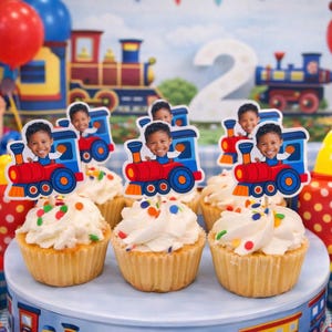 May include: Cupcakes with white icing and colourful sprinkles, decorated with train-shaped toppers featuring a child's face. The background includes a train-themed backdrop and balloons, ideal for a birthday party.