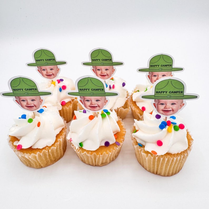 Op de afbeelding: Zeven cupcakes met witte glazuur en kleurrijke hagelslag. Elke cupcake is bekroond met een papieren uitsparing van een baby met een groene hoed met de tekst "Happy Camper". De cupcakes staan op een wit oppervlak.