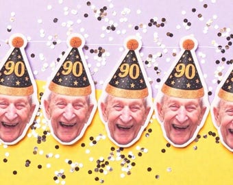 Puede incluir: Una guirnalda de decoraciones de fiesta con una persona mayor sonriente que lleva un gorro de fiesta negro y dorado con el número 90. El fondo es amarillo y morado con confeti disperso.