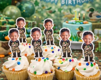 Puede incluir: Cupcakes con glaseado blanco y chispas de colores, cubiertos con recortes de un niño sonriente con monos de camuflaje. Una pancarta que dice "Happy Birthday" está en el fondo, junto con un pastel decorado y globos.