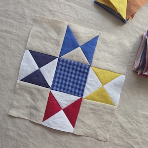 May include: A square quilt block with a star design. The center is a blue and white gingham pattern, surrounded by blue, yellow, red, and white triangles on a beige background. Additional fabric squares are visible in the background.