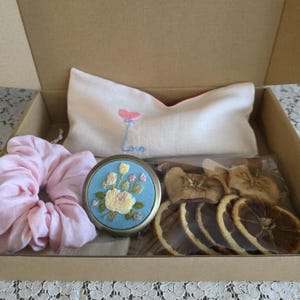 May include: A gift box containing a pink scrunchie, a compact mirror with floral embroidery, a white pillow with a pink balloon and the word "Love", and dried fruit slices. The box is made of cardboard.