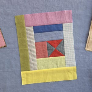 May include: A rectangular fabric quilt block featuring a patchwork design of various colors and patterns. The block includes strips of solid olive green, pink, blue, light green, and yellow fabric, alongside sections of white and blue gingham.