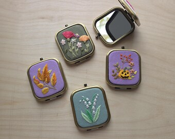 May include: Five square compact mirrors with brass-toned metal frames. Each features a unique embroidered fabric cover: one with autumn leaves, one with a jack-o'-lantern, one with lily of the valley, one with wildflowers, and one open mirror.