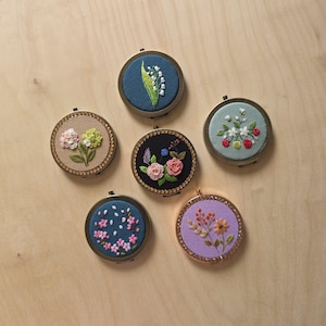 May include: Six round compact mirrors featuring embroidered fabric tops. Designs include lily of the valley on dark blue, hydrangea on beige, a floral bouquet on black, strawberries on light blue, cherry blossoms on blue, and wildflowers on purple.