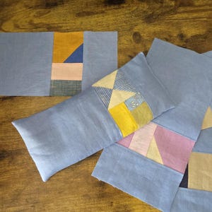 May include: Several light blue fabric pieces with patchwork designs. The patchwork includes geometric shapes in yellow, pink, blue, and green. The pieces are arranged on a wooden surface, showcasing the handmade quality.