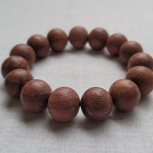 May include: A bracelet composed of fourteen round, wooden beads. The beads are a warm, reddish-brown color and appear to be strung together to form a circular shape. The bracelet is displayed on a light-colored, textured surface.