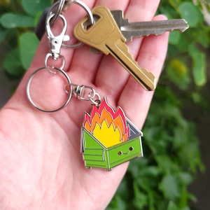 Peut inclure: Un porte-clés en métal vert en forme de benne à ordures avec des flammes qui sortent du haut. La benne à ordures a un visage souriant sur le devant. Le porte-clés est attaché à un porte-clés argenté avec deux clés.