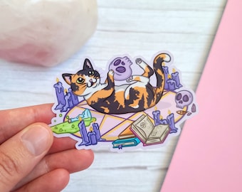 May include: A calico cat with green eyes is lying on a pentagram with a purple skull in its paws. There are purple candles, a green potion bottle, and two books on the pentagram.