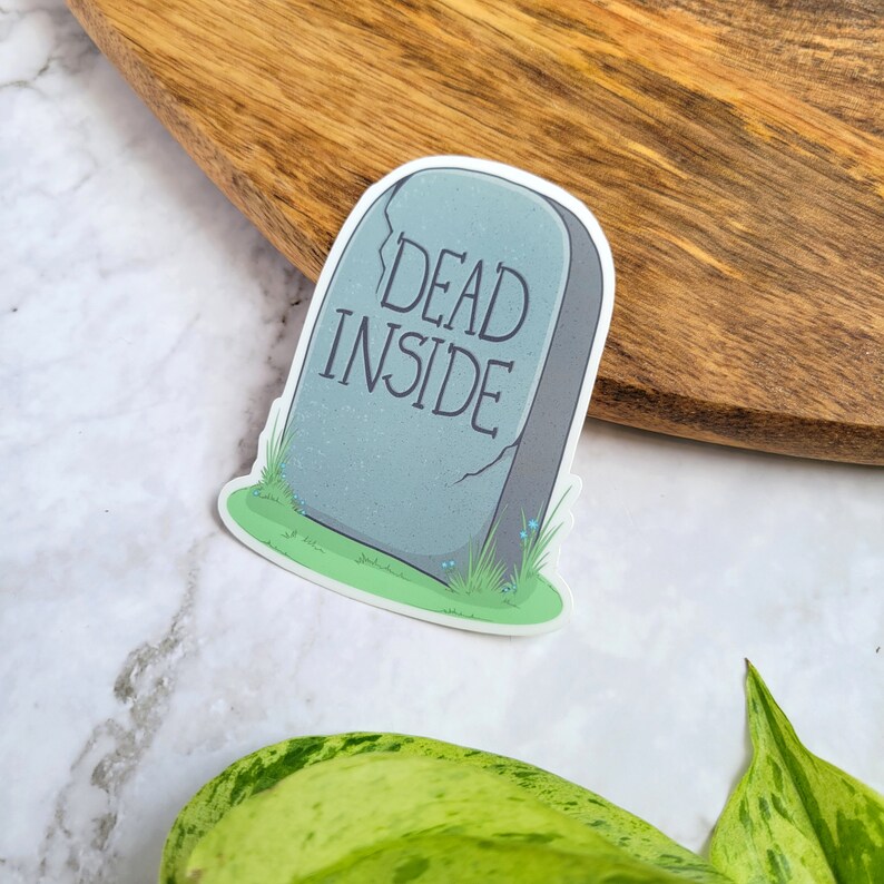 May include: A gray tombstone sticker with the text "DEAD INSIDE" in black lettering. The sticker is surrounded by green grass and blue flowers.
