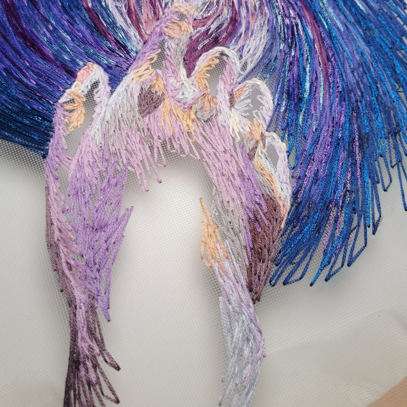 May include: Close-up of a purple, blue, and white embroidered design on a white fabric background. The embroidery is detailed and intricate, with a variety of stitches and textures.