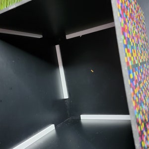 May include: A black cube-shaped box with a colourful pixelated pattern on one side. The inside of the box is black with white trim around the edges.