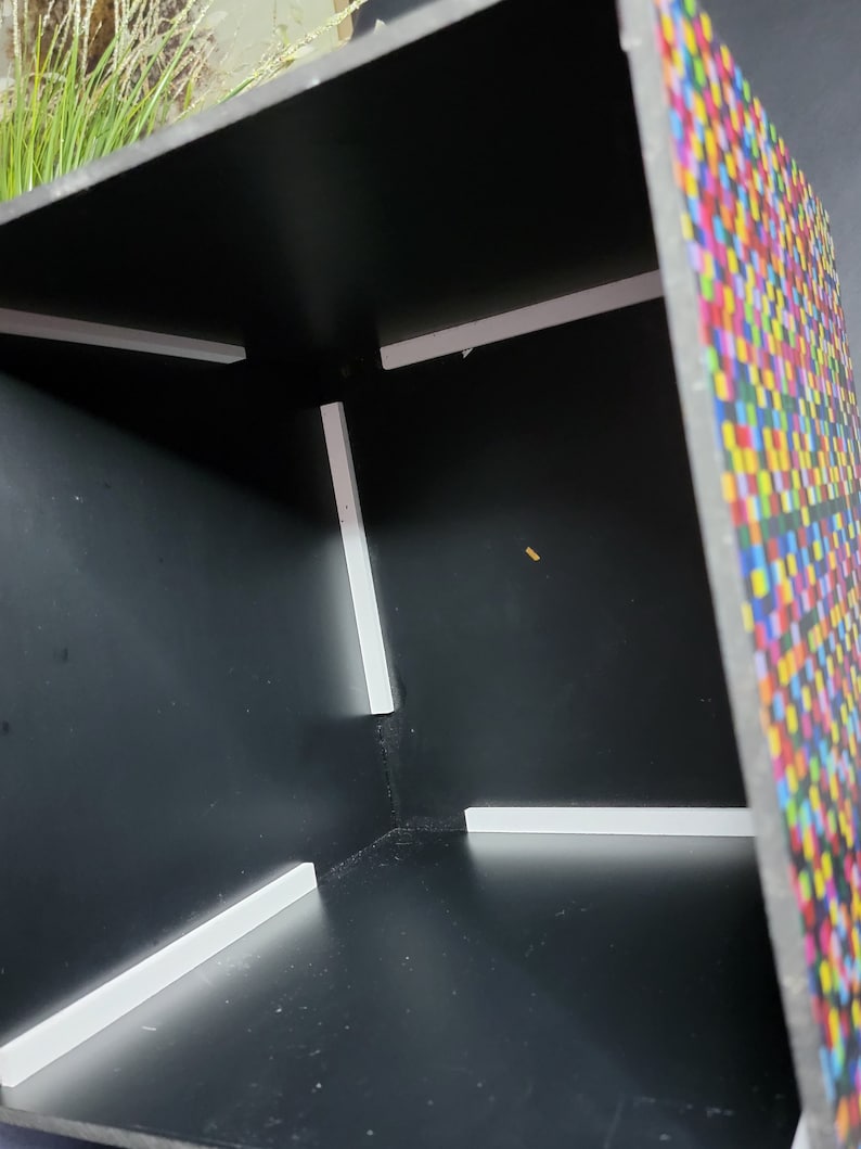 May include: A black cube-shaped box with a colourful pixelated pattern on one side. The inside of the box is black with white trim around the edges.