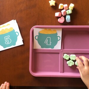 May include: A pink tray with four compartments.  The top left compartment has a card with a green pot of gold with the number 4 on it. The top right compartment has a card with a green pot of gold with the number 5 on it. The bottom right compartment has five green shamrock-shaped marshmallows.