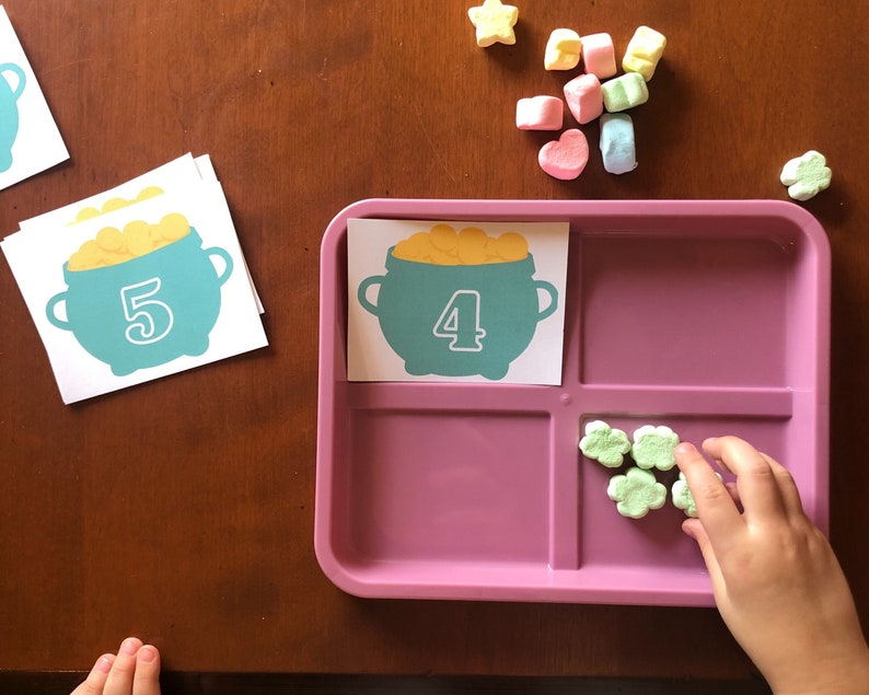 May include: A pink tray with four compartments.  The top left compartment has a card with a green pot of gold with the number 4 on it. The top right compartment has a card with a green pot of gold with the number 5 on it. The bottom right compartment has five green shamrock-shaped marshmallows.