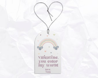 Op de afbeelding: Een wit cadeau-etiket met een regenboog, twee wolken met smiley-gezichten en roze hartjes. De tekst op het etiket luidt "Valentine, jij kleurt mijn wereld. Liefs, Nora."