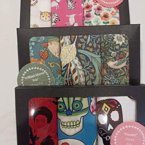 以下が含まれることがあります： 3つのファットクォーター生地のセット。上段の箱は猫柄、中段の箱はウィリアム・モリスのデザイン、下段の箱はフリーダ・カーロとスカル柄です。各箱には丸いステッカーが貼られています。