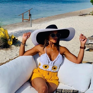May include: A woman wearing a white crochet sunflower top, yellow shorts, and a wide-brimmed black and white sun hat. She is sitting on a white couch with a black and white striped clutch.