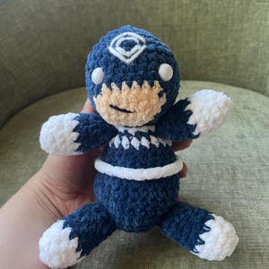 May include: A handmade blue and white crochet doll with a smiling face. The doll has white accents on its arms and feet, and a white band around its waist. The doll's head features a white diamond-shaped design.