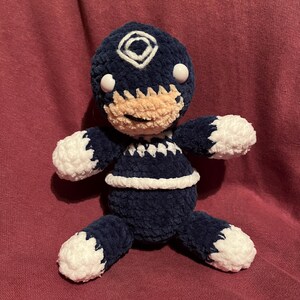 May include: A handmade plush doll in navy blue and white. The doll has a round head with a white diamond-shaped design and two white eyes. The doll's face is tan, and it has white accents on the arms, legs, and torso. The doll is approximately 8 inches tall.