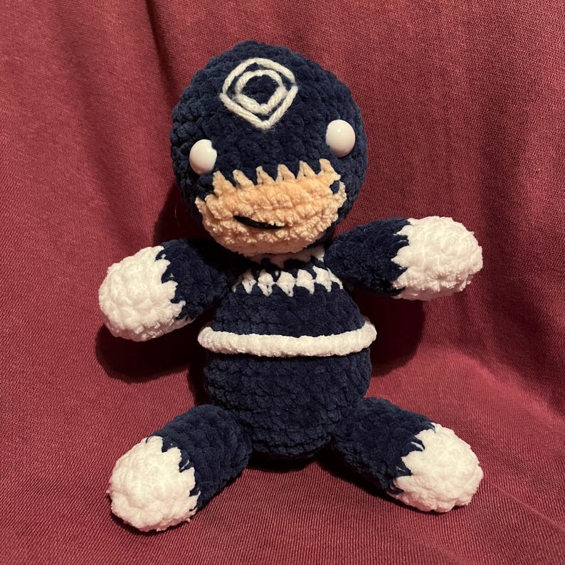 May include: A handmade plush doll in navy blue and white. The doll has a round head with a white diamond-shaped design and two white eyes. The doll's face is tan, and it has white accents on the arms, legs, and torso. The doll is approximately 8 inches tall.