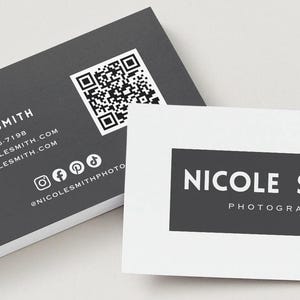 Op de afbeelding: Twee rechthoekige visitekaartjes in een zwart-wit kleurenschema. Eén kaart toont de naam Nicole Smith, een QR-code, telefoonnummer, e-mailadres, website en social media iconen. De andere kaart bevat de tekst Nicole Smith Photography.