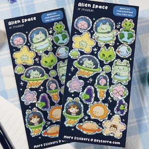 May include: Two sticker sheets with alien-themed designs. Each sheet features various stickers of aliens in spaceships, planets, and stars. The text "Alien Space" and "35 Stickers" is visible. The stickers have a holographic finish. A toy spaceship and washi tape are also in the image.