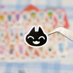 May include: A black and white sticker of a smiling cat face with a simple design. The sticker is being held by tweezers.