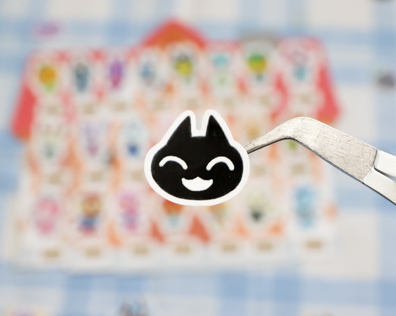 May include: A black and white sticker of a smiling cat face with a simple design. The sticker is being held by tweezers.