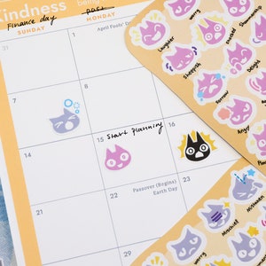 May include: A monthly planner with a yellow and white cover. The planner has a calendar grid with the days of the week labeled. The planner also has a section for "Start Planning" and a section for "Passover (Begins) Earth Day". The planner is decorated with colorful cat stickers with different emotions.