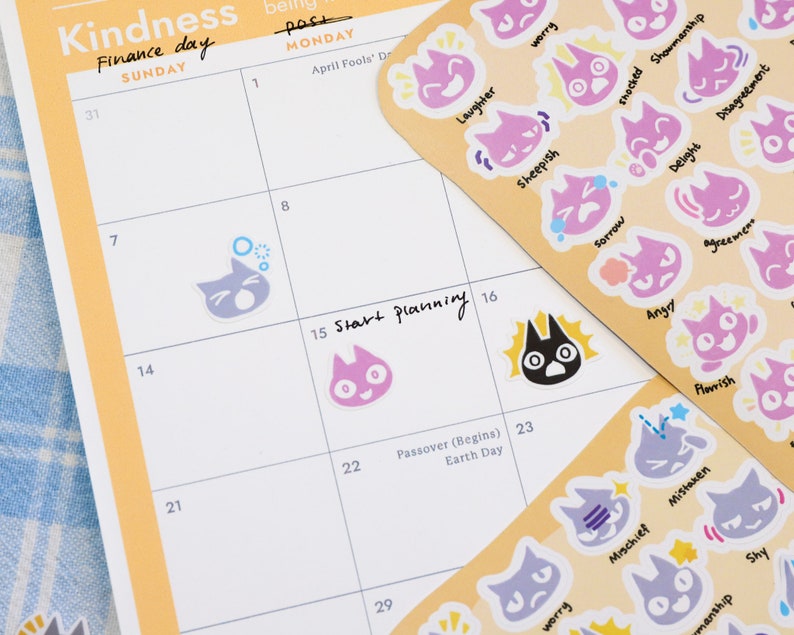 May include: A monthly planner with a yellow and white cover. The planner has a calendar grid with the days of the week labeled. The planner also has a section for "Start Planning" and a section for "Passover (Begins) Earth Day". The planner is decorated with colorful cat stickers with different emotions.