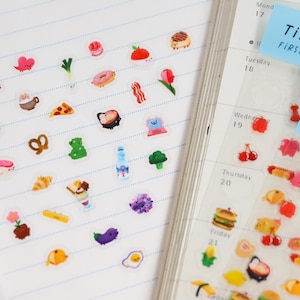 May include: A sheet of clear stickers with colorful illustrations of food, drinks, and other objects. The stickers are arranged in rows on a white notebook page. The text "TINY TOWN FIRST EDITION" is visible on a blue sticker.
