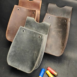 Op de afbeelding: Drie leren tasjes in tinten bruin, donkerbruin en antracietgrijs, elk met een gebogen bovenrand, zichtbare stiksels en twee metalen klinknagels. Drie jachtpatronen in blauw, geel en rood zijn voor de tasjes geplaatst.
