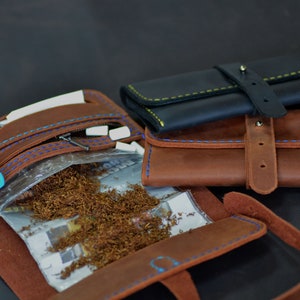 May include: A brown leather tobacco pouch with a zipped pocket, a clear plastic bag containing loose tobacco, a blue lighter, and white paper. The pouch has blue stitching detail and a brown leather strap with a buckle.