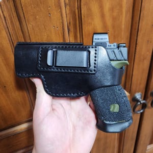 Puede incluir: Funda de cuero negro con clip metálico, que sujeta una pistola negra y verde con mira de punto rojo. La empuñadura de la pistola tiene una superficie texturizada.
