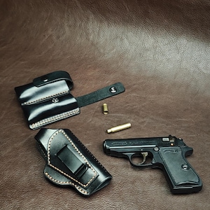 Op de afbeelding: Zwarte leren holster- en magazijntasjeset met een zwart handvuurwapen en twee kogels. De holster heeft een riemclip. Het handvuurwapen is zwart met het woord "Walther" aan de zijkant. De achtergrond is bruin.