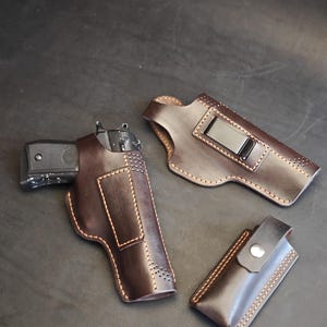 Puede incluir: Un conjunto de fundas de cuero marrón y una funda para cargador. Una pistola negra está en una funda. El cuero está cosido con hilo marrón claro. Las fundas tienen un clip para sujetar al cinturón.