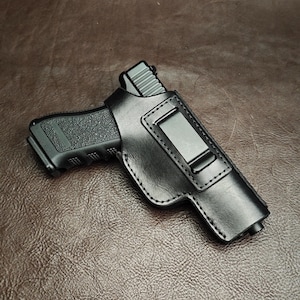 Puede incluir: Una funda de cuero negro que contiene una pistola semiautomática negra. La funda presenta un acabado liso, costuras visibles a lo largo de los bordes y un clip de cinturón de metal resistente fijado en la parte frontal para sujeción.