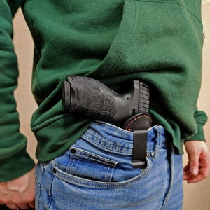 Puede incluir: Una pistola negra con una funda de cuero marrón está metida en la cintura de un pantalón vaquero azul.