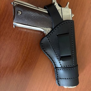 Puede incluir: Una pistola semiautomática de color plateado con empuñadura texturizada oscura está parcialmente insertada en una funda de cuero negro. La funda presenta costuras negras visibles, un clip de cinturón de metal y una correa de sujeción.
