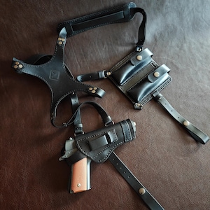 Op de afbeelding: Een zwart lederen schouderholstersysteem met een handvuurwapen met bruine greep, twee magazijntasjes met drukknoopsluitingen, verstelbare riemen, metalen klinknagels en een bevestigingsriem voor aan de broekriem.