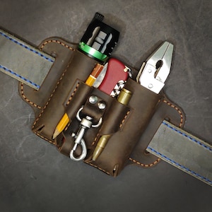 Puede incluir: Un cinturón de herramientas de cuero marrón con múltiples bolsillos y bucles. El cinturón tiene un detalle de costura azul. Los bolsillos contienen una linterna, una multiherramienta, un lápiz, un par de alicates y un cuchillo pequeño.