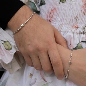Puede incluir: Primer plano de dos muñecas, cada una adornada con una delicada pulsera de plata. Las pulseras presentan pequeñas cuentas y dijes en forma de corazón. El fondo muestra un vestido con estampado floral y una manga negra.