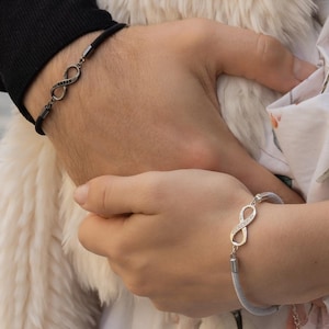 Puede incluir: Dos pulseras con el símbolo del infinito. Una es negra, la otra plateada. Las pulseras se llevan en las muñecas. El fondo incluye un chal cremoso y esponjoso y un vestido con estampado floral.